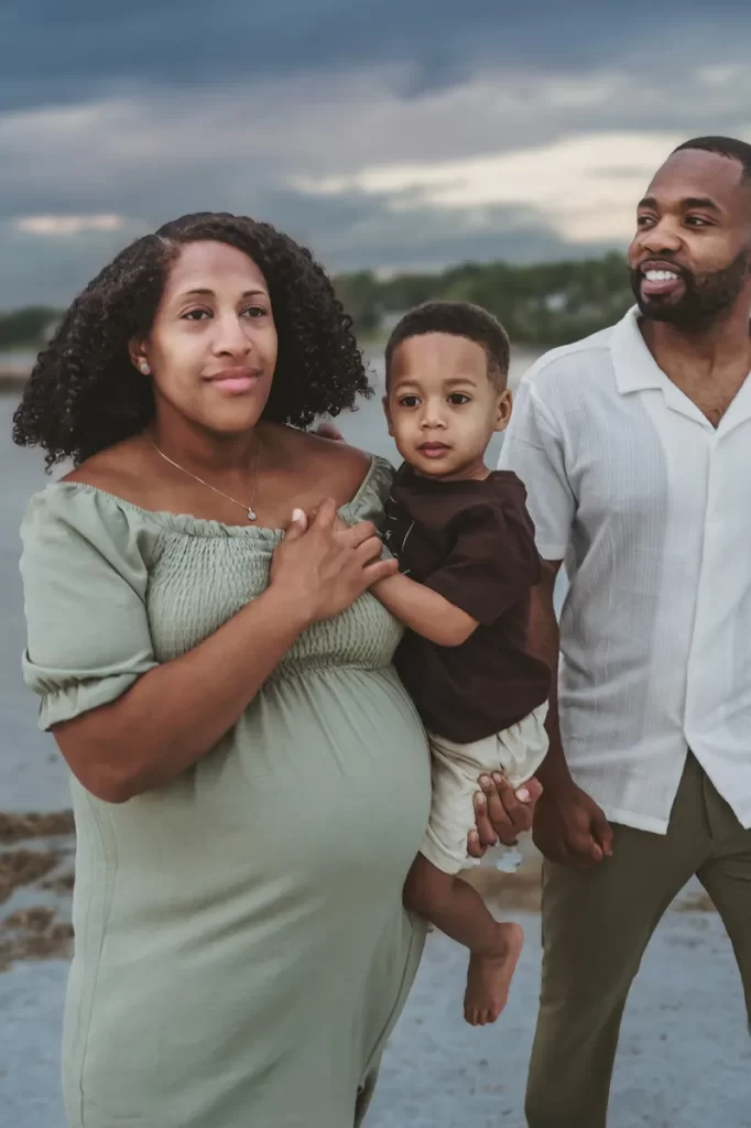 A little boy being held by his pregnant mom walking with his dadas they stand on the beach at sunset on an overcast evening