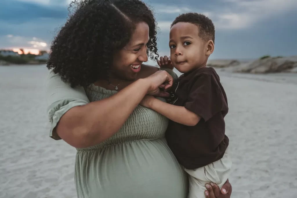 A little boy being held by his pregnant mom as they stand on the beach at sunset on an overcast evening